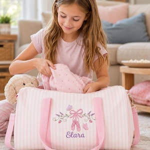 May include: A light pink and white striped duffel bag featuring an embroidered design of ballet slippers, a bow, flowers, and butterflies. The name Elara is embroidered in purple script below the ballet slippers. The bag has pink handles.