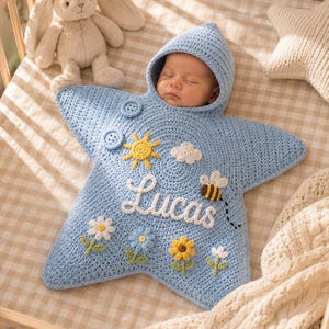 May include: A light blue crocheted star-shaped baby swaddle with a hood, featuring the name Lucas embroidered in white script. The design includes a yellow sun, a white cloud, a small bee, and four colorful flowers, with two buttons at the top.