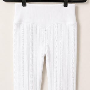 May include: High-waisted white leggings featuring a vertical ribbed texture and a subtle cable knit pattern. The garment has a wide, elasticated waistband and is suspended from a black plastic hanger with two clips at the top edge.