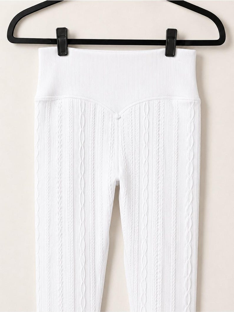 May include: High-waisted white leggings featuring a vertical ribbed texture and a subtle cable knit pattern. The garment has a wide, elasticated waistband and is suspended from a black plastic hanger with two clips at the top edge.