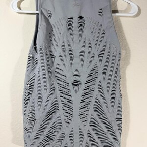 May include: A light gray sleeveless tank top featuring an intricate, laser-cut pattern of horizontal slits across the entire front. The garment has a round neckline and displays the brand logo 'alo' printed in small letters near the collar.