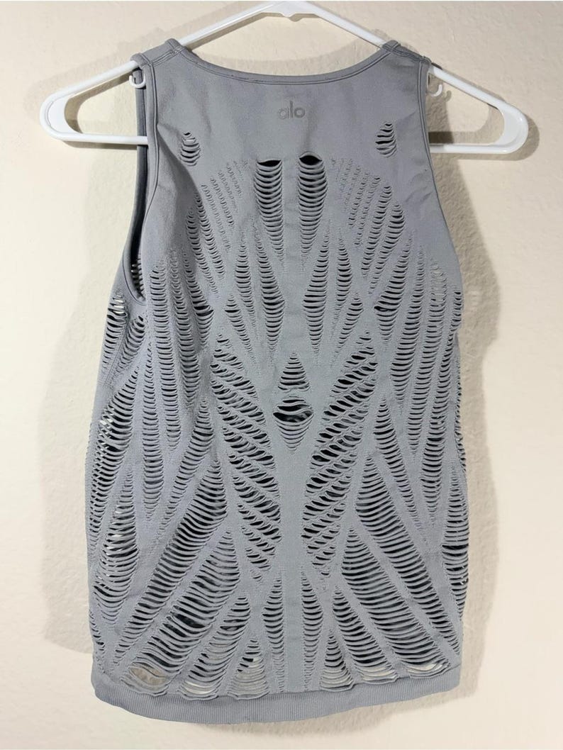 May include: A light gray sleeveless tank top featuring an intricate, laser-cut pattern of horizontal slits across the entire front. The garment has a round neckline and displays the brand logo 'alo' printed in small letters near the collar.