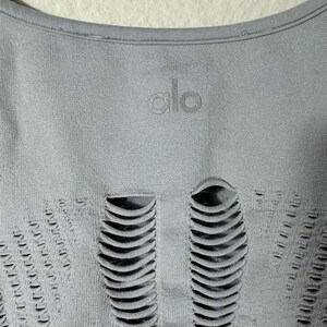 May include: A light gray athletic top featuring a round neckline and the word alo printed in a matching gray tone. The fabric displays a series of vertical, elongated cut-out details arranged in a symmetrical pattern across the front surface.