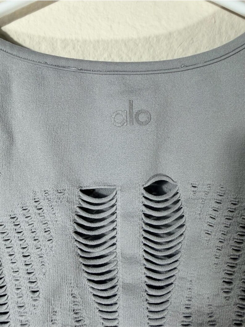 May include: A light gray athletic top featuring a round neckline and the word alo printed in a matching gray tone. The fabric displays a series of vertical, elongated cut-out details arranged in a symmetrical pattern across the front surface.