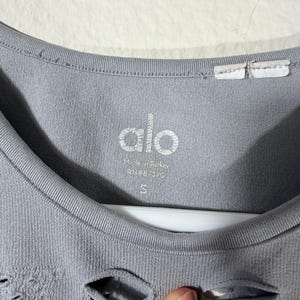 May include: A light gray Alo brand garment featuring a round neckline and a textured, distressed pattern with small cutouts. The interior label displays the text Alo, Made in Turkey, RN#87370, and the size S, printed in white on the fabric.