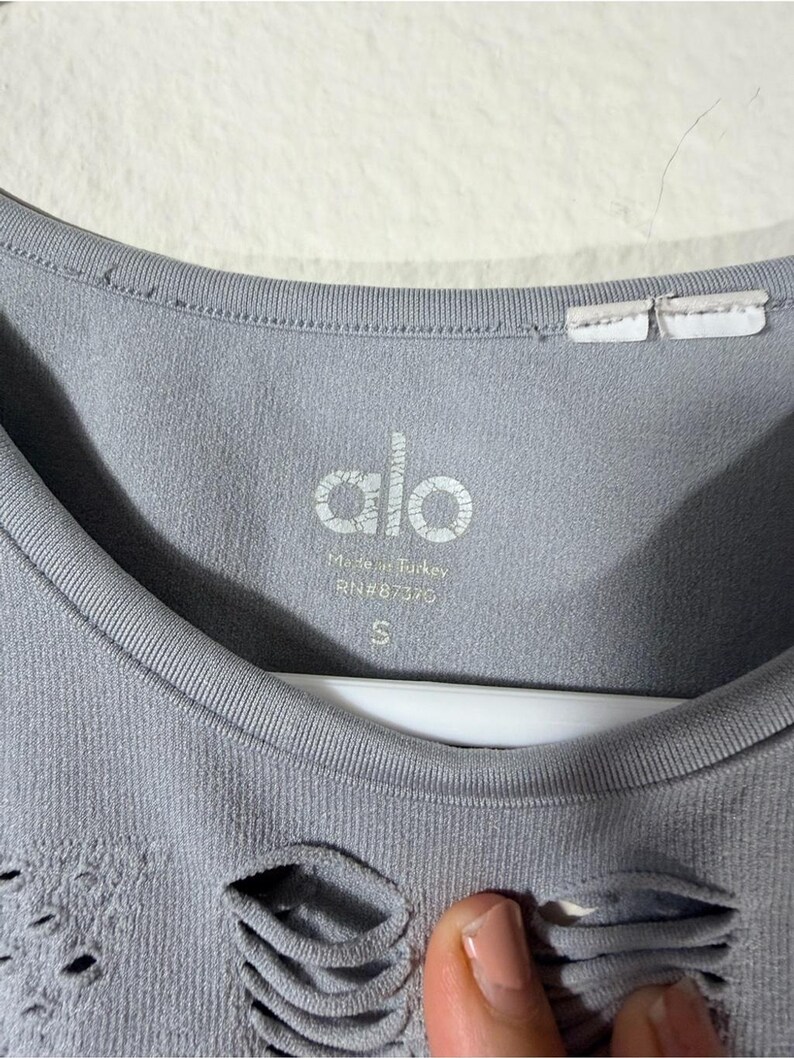 May include: A light gray Alo brand garment featuring a round neckline and a textured, distressed pattern with small cutouts. The interior label displays the text Alo, Made in Turkey, RN#87370, and the size S, printed in white on the fabric.