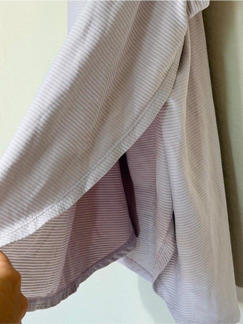 Peut inclure: Un v&ecirc;tement lilas clair pr&eacute;sentant une texture finement c&ocirc;tel&eacute;e et une fente lat&eacute;rale. Le tissu affiche un motif &agrave; rayures verticales coh&eacute;rent sur toute la surface, avec des coutures visibles le long de l'ourlet et de l'ouverture lat&eacute;rale.