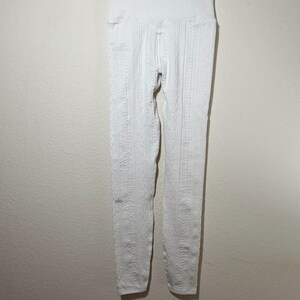 May include: A pair of white high-waisted leggings featuring a vertical cable knit texture throughout the fabric. The leggings have a wide, ribbed waistband and a small, subtle logo printed in silver on the left leg near the lower calf area.