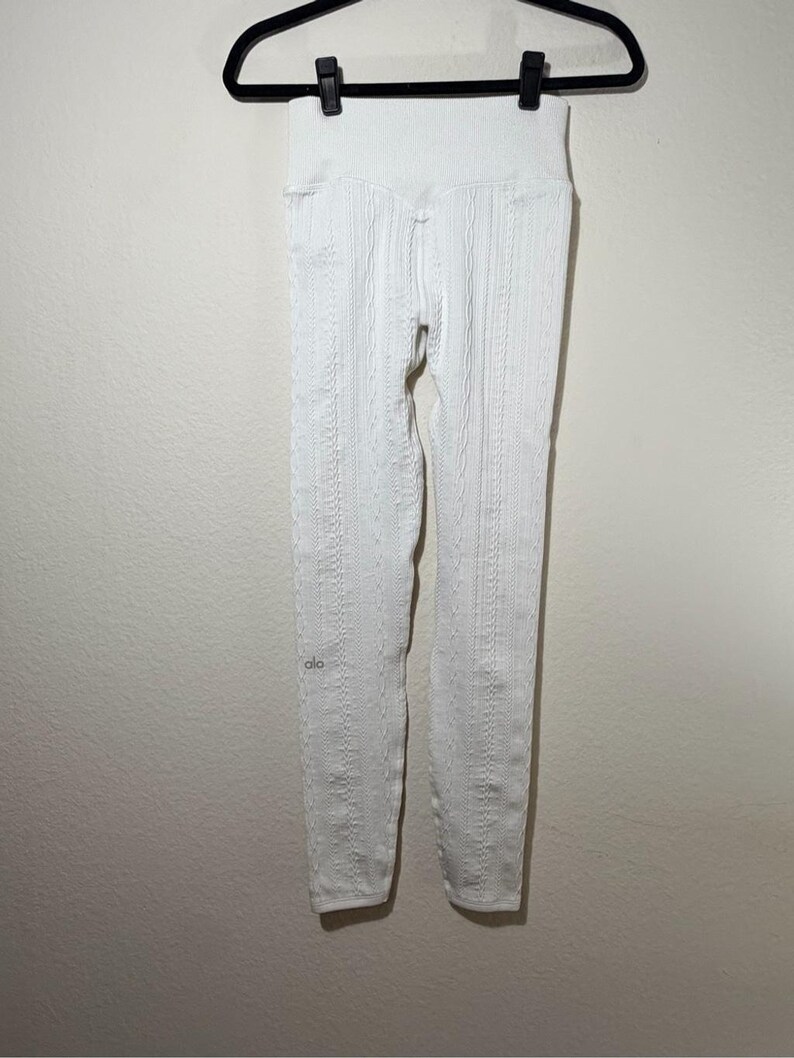 May include: A pair of white high-waisted leggings featuring a vertical cable knit texture throughout the fabric. The leggings have a wide, ribbed waistband and a small, subtle logo printed in silver on the left leg near the lower calf area.