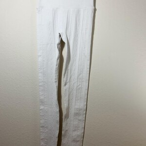 May include: A pair of white leggings featuring a vertical cable knit pattern and a wide, ribbed high-waist band. The leggings are constructed from a textured fabric and are displayed hanging from a black plastic clothes hanger.
