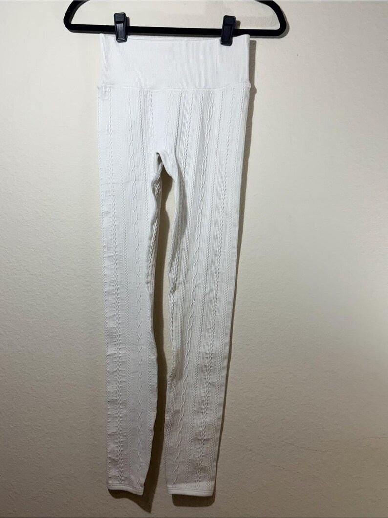 May include: A pair of white leggings featuring a vertical cable knit pattern and a wide, ribbed high-waist band. The leggings are constructed from a textured fabric and are displayed hanging from a black plastic clothes hanger.