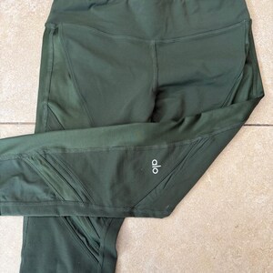 Peut inclure: Legging vert olive fonc&eacute; dot&eacute; d'une taille haute et de panneaux en maille transparente sur les c&ocirc;t&eacute;s. Le v&ecirc;tement pr&eacute;sente des coutures visibles et le texte blanc "alo" imprim&eacute; sur la partie inf&eacute;rieure de la jambe gauche du pantalon.