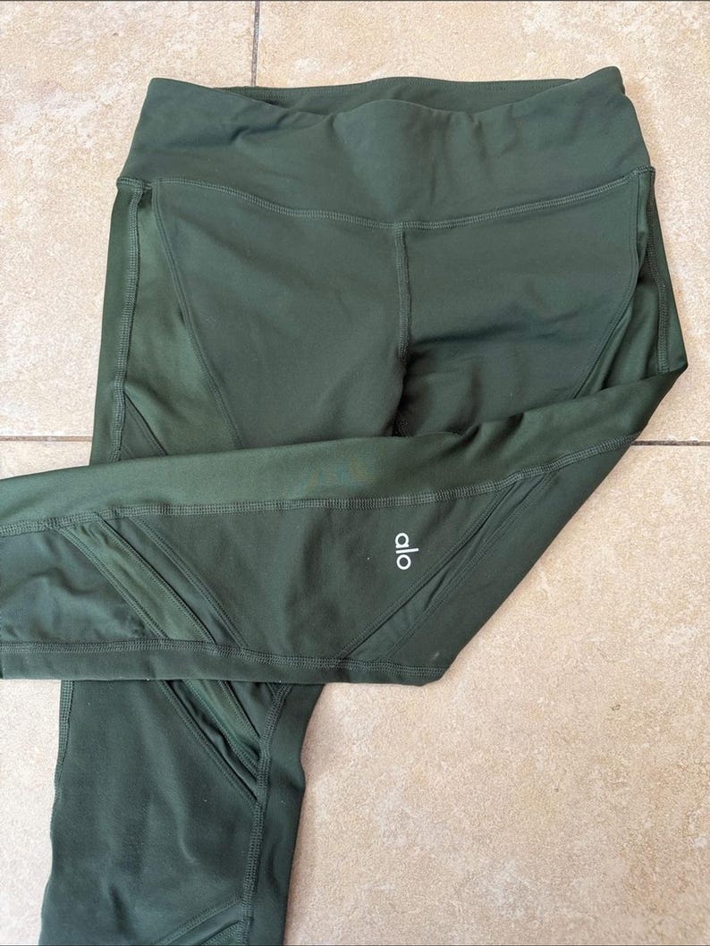 Peut inclure: Legging vert olive fonc&eacute; dot&eacute; d'une taille haute et de panneaux en maille transparente sur les c&ocirc;t&eacute;s. Le v&ecirc;tement pr&eacute;sente des coutures visibles et le texte blanc "alo" imprim&eacute; sur la partie inf&eacute;rieure de la jambe gauche du pantalon.