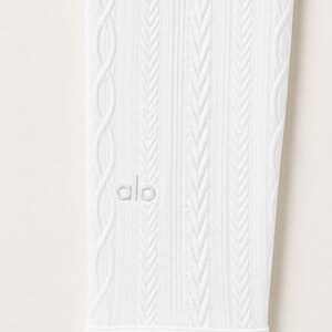 May include: A white legging featuring a vertical cable knit texture and ribbed detailing. The fabric displays a consistent pattern of raised braided lines running the length of the garment. The word alo is embroidered in white on the lower section.