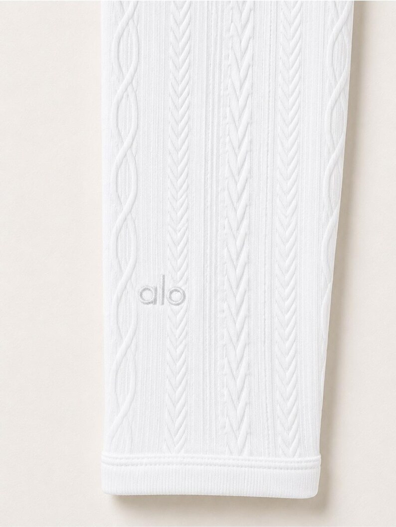 May include: A white legging featuring a vertical cable knit texture and ribbed detailing. The fabric displays a consistent pattern of raised braided lines running the length of the garment. The word alo is embroidered in white on the lower section.