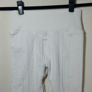 May include: A pair of white leggings featuring a high-waisted ribbed waistband and a vertical cable knit texture pattern throughout the fabric. The garment is suspended from a black plastic hanger with two clips securing the top edge of the waistband.