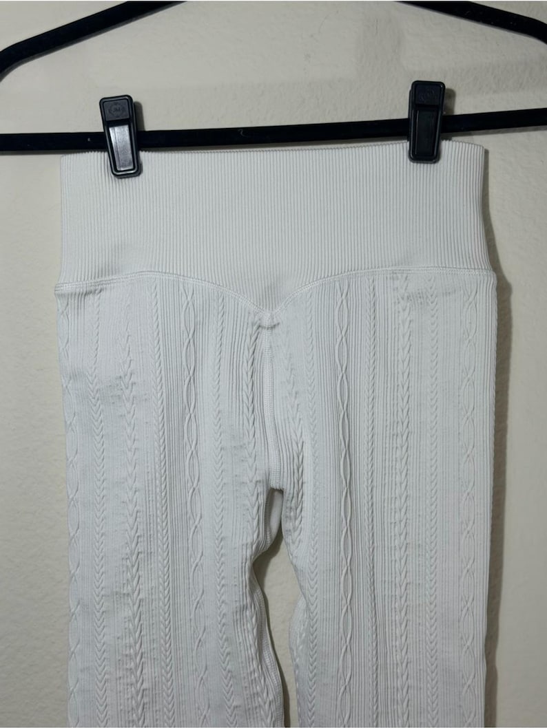 May include: A pair of white leggings featuring a high-waisted ribbed waistband and a vertical cable knit texture pattern throughout the fabric. The garment is suspended from a black plastic hanger with two clips securing the top edge of the waistband.