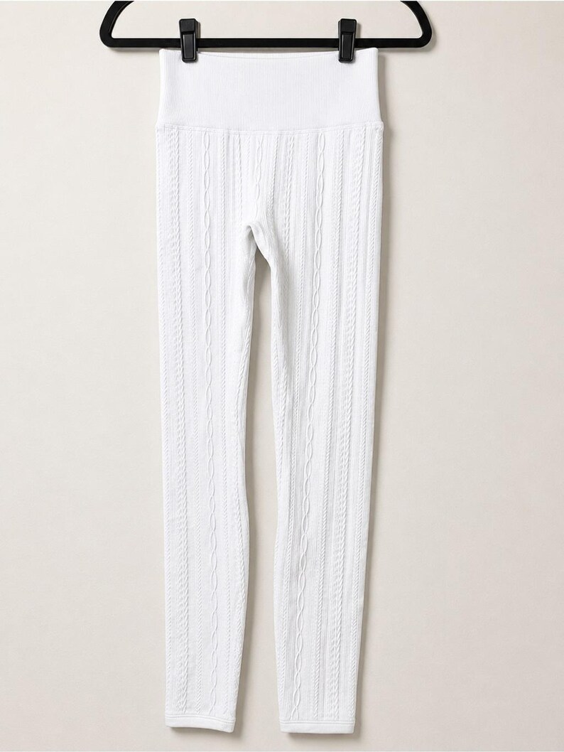 May include: A pair of white leggings featuring a vertical cable knit pattern throughout the legs and a wide, ribbed high-waist band. The leggings are displayed hanging from a black plastic hanger against a plain, light-coloured background.