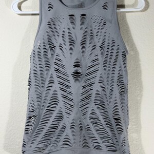 May include: A grey sleeveless tank top featuring an intricate, all-over laser-cut pattern of horizontal slits. The garment has a round neckline, a ribbed hem, and the brand name Alo is printed in small letters on the inside of the neck area.
