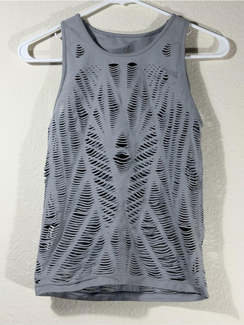 May include: A grey sleeveless tank top featuring an intricate, all-over laser-cut pattern of horizontal slits. The garment has a round neckline, a ribbed hem, and the brand name Alo is printed in small letters on the inside of the neck area.