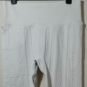 May include: Off-white leggings featuring a high-rise waistband and a vertical cable knit texture pattern. The garment is constructed with a ribbed fabric design that runs throughout the length of the legs, providing a consistent and uniform appearance.