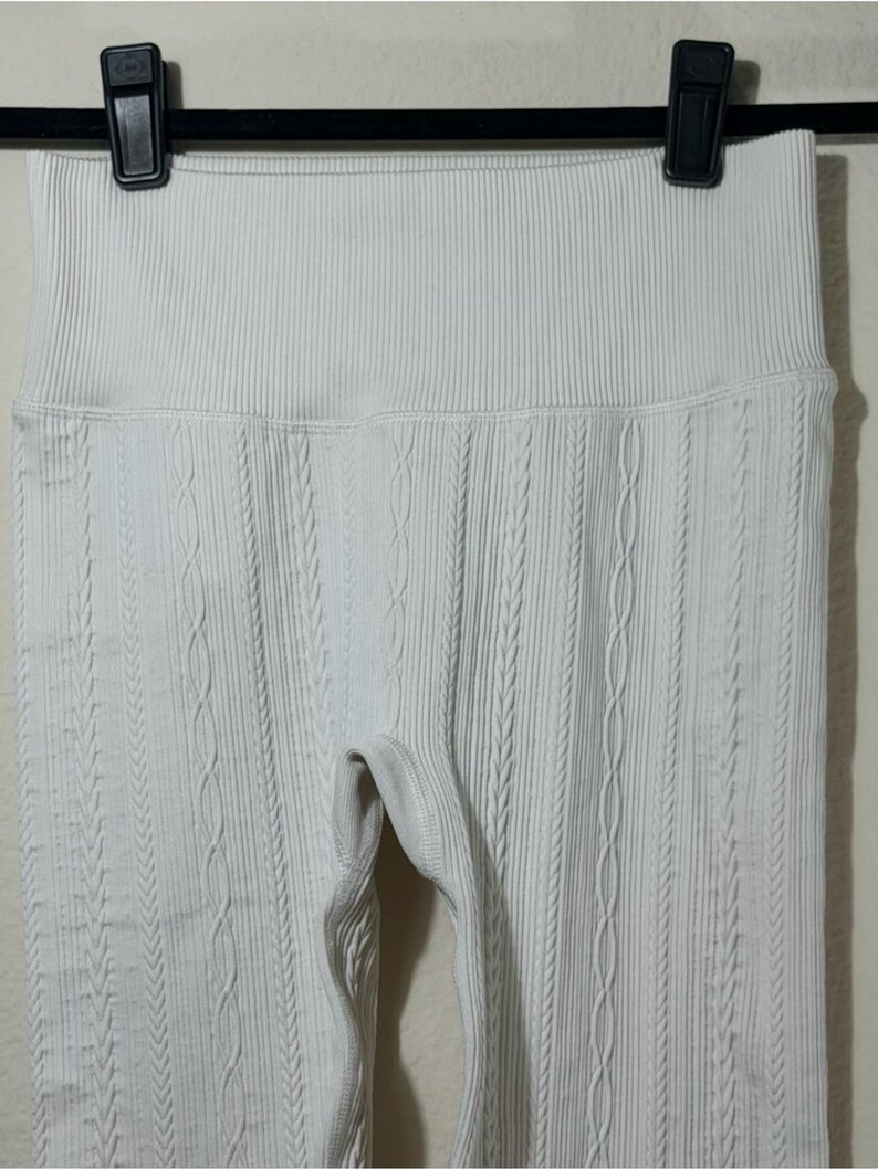May include: Off-white leggings featuring a high-rise waistband and a vertical cable knit texture pattern. The garment is constructed with a ribbed fabric design that runs throughout the length of the legs, providing a consistent and uniform appearance.