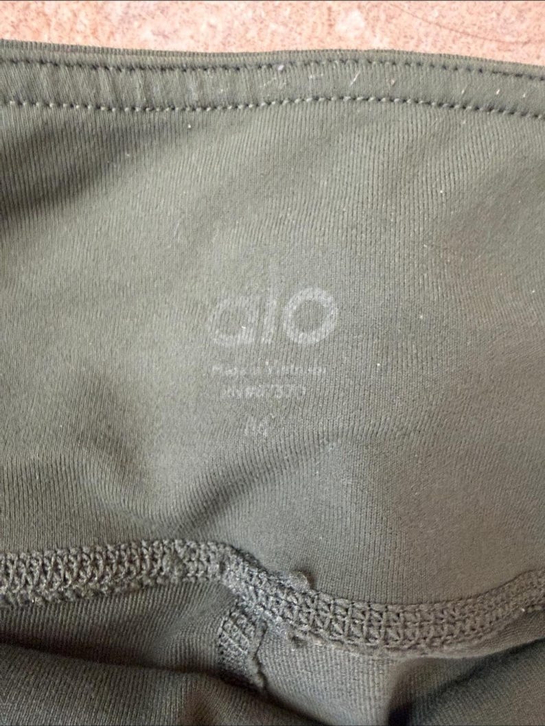 Peut inclure: Un legging vert olive dot&eacute; d'une taille haute avec le nom de la marque alo imprim&eacute; en blanc. Sous le nom de la marque, le texte indique Made in Vietnam, RN# 107370, et la taille M est clairement imprim&eacute;e &agrave; l'int&eacute;rieur du tissu.