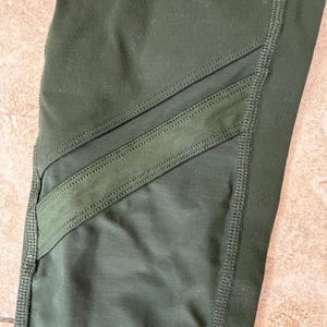 Peut inclure: Legging vert olive dot&eacute; de deux empi&egrave;cements en maille filet diagonaux sur la zone de la cuisse. Le v&ecirc;tement est confectionn&eacute; avec des coutures renforc&eacute;es visibles le long des jointures et des bords des inserts en maille, cr&eacute;ant un aspect textur&eacute;.