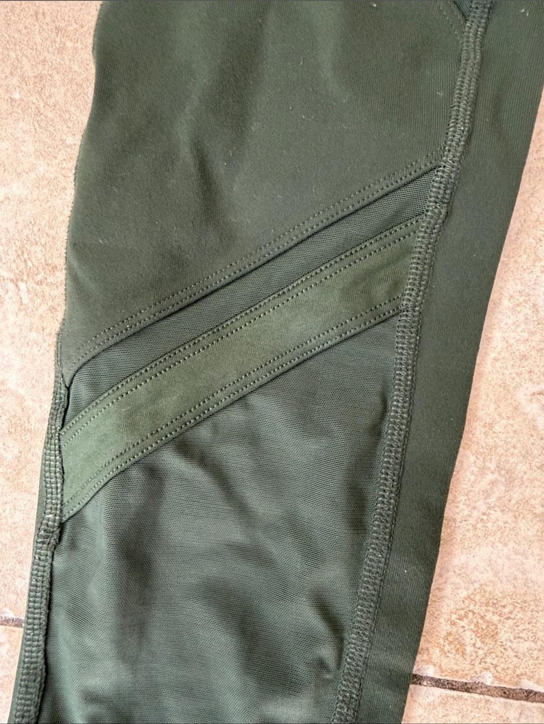Peut inclure: Legging vert olive dot&eacute; de deux empi&egrave;cements en maille filet diagonaux sur la zone de la cuisse. Le v&ecirc;tement est confectionn&eacute; avec des coutures renforc&eacute;es visibles le long des jointures et des bords des inserts en maille, cr&eacute;ant un aspect textur&eacute;.