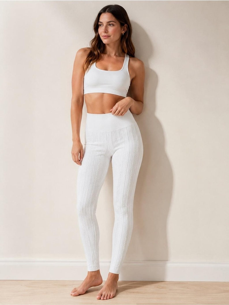 May include: A white two-piece athletic set consisting of a scoop-neck sports bra and high-waisted leggings. The leggings feature a vertical cable-knit texture throughout, while the sports bra has a smooth, solid finish with a wide under-bust band.