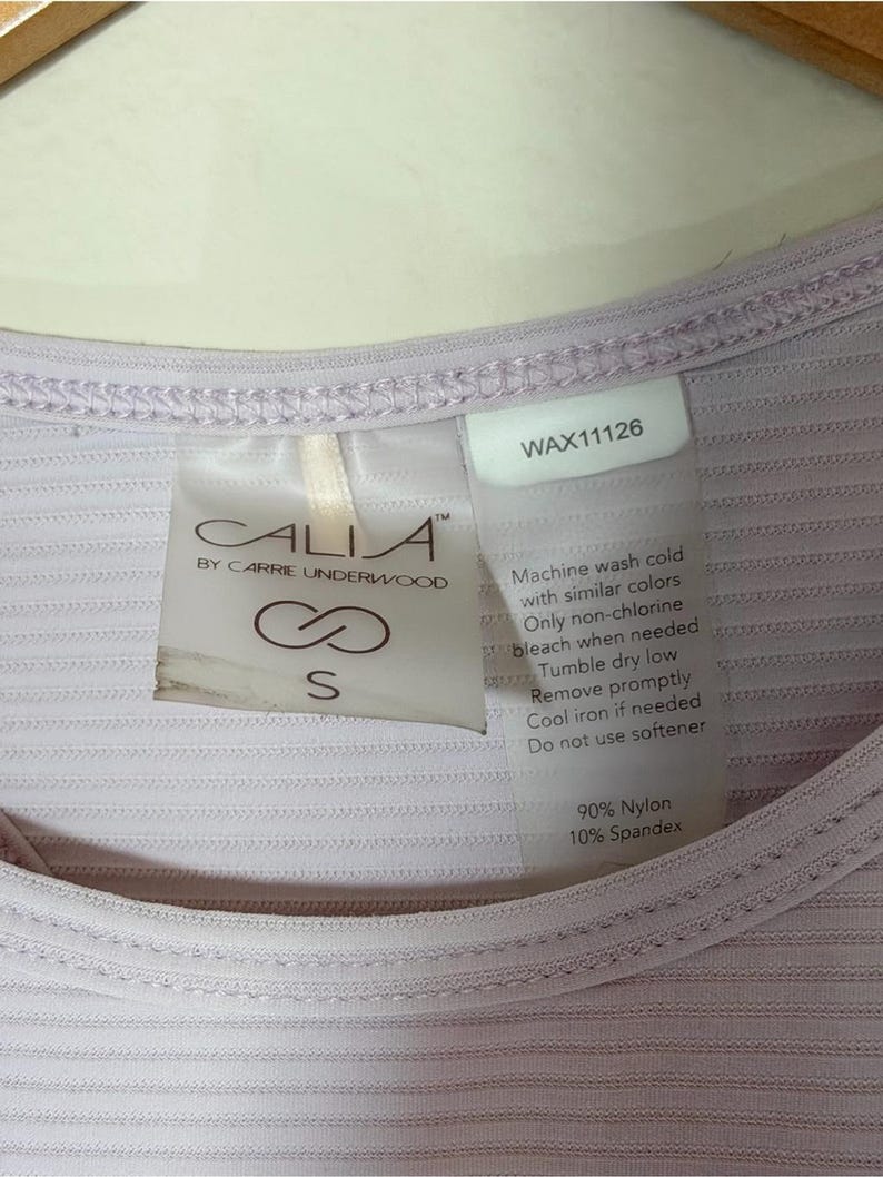 Peut inclure: Un haut c&ocirc;tel&eacute; lilas clair avec une &eacute;tiquette de marque Calia by Carrie Underwood, taille S, et une &eacute;tiquette d'entretien. Le v&ecirc;tement est compos&eacute; de 90 % de nylon et 10 % d'&eacute;lasthanne, avec le texte WAX11126 et des instructions de lavage.