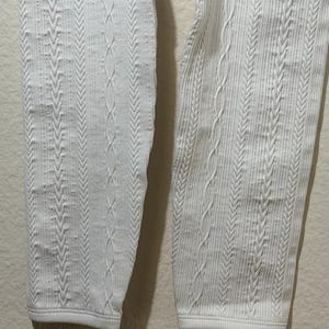 May include: A pair of white leggings featuring a vertical cable knit pattern. The fabric displays a textured design with alternating columns of braided cable stitches and fine ribbed lines, extending down to the straight, hemmed leg openings.