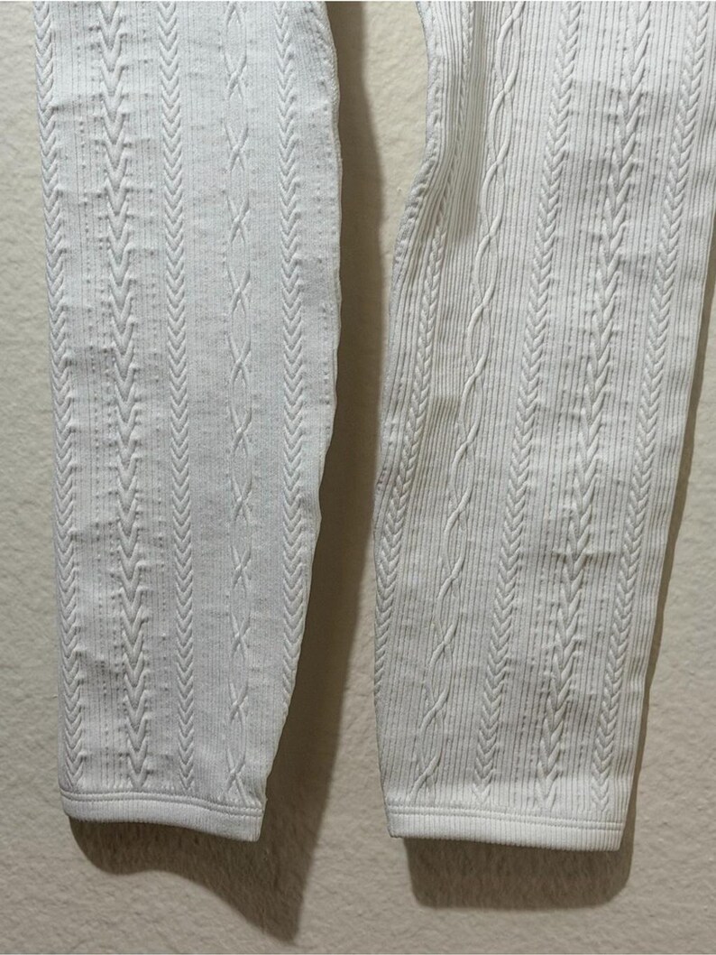 May include: A pair of white leggings featuring a vertical cable knit pattern. The fabric displays a textured design with alternating columns of braided cable stitches and fine ribbed lines, extending down to the straight, hemmed leg openings.