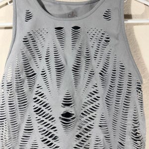 May include: A light gray sleeveless tank top featuring a complex, laser-cut pattern of horizontal slits across the front. The garment has a round neckline and is displayed on a white hanger, revealing a dark layer underneath the cutouts.