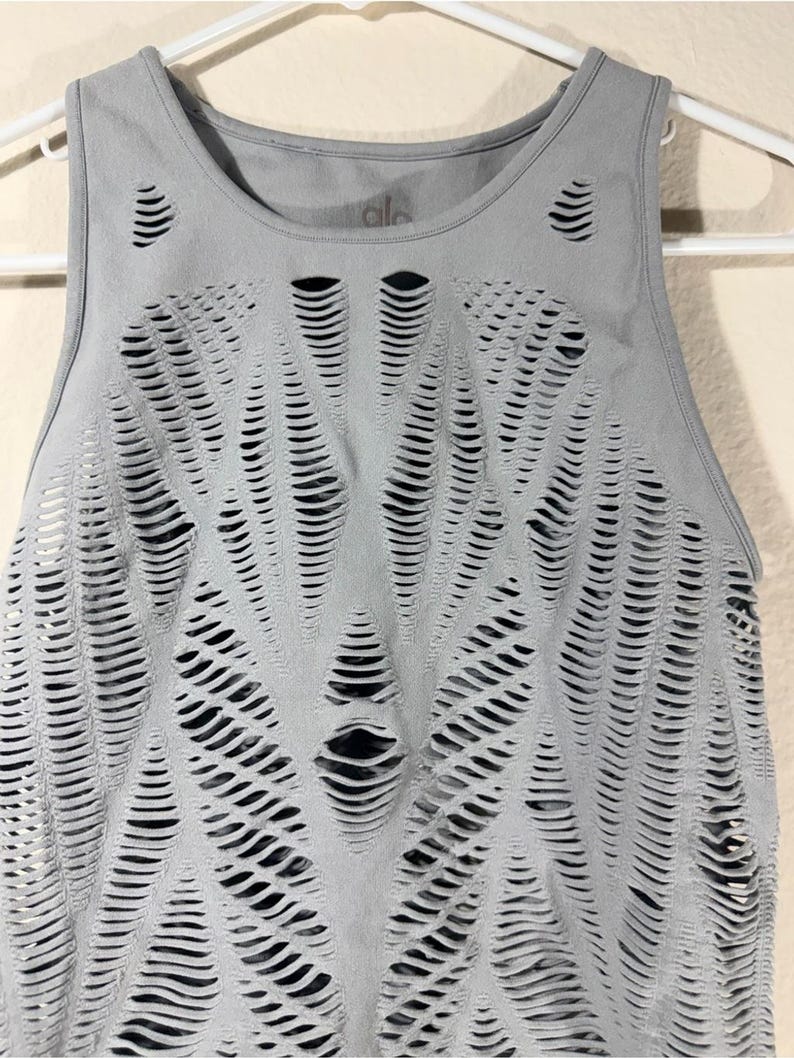 May include: A light gray sleeveless tank top featuring a complex, laser-cut pattern of horizontal slits across the front. The garment has a round neckline and is displayed on a white hanger, revealing a dark layer underneath the cutouts.