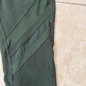 Peut inclure: Legging vert olive fonc&eacute; dot&eacute; de trois panneaux en maille diagonaux sur le c&ocirc;t&eacute;. Le v&ecirc;tement est con&ccedil;u avec des coutures renforc&eacute;es visibles le long des jointures et des bords des inserts en maille transparente, cr&eacute;ant un aspect athl&eacute;tique structur&eacute;.