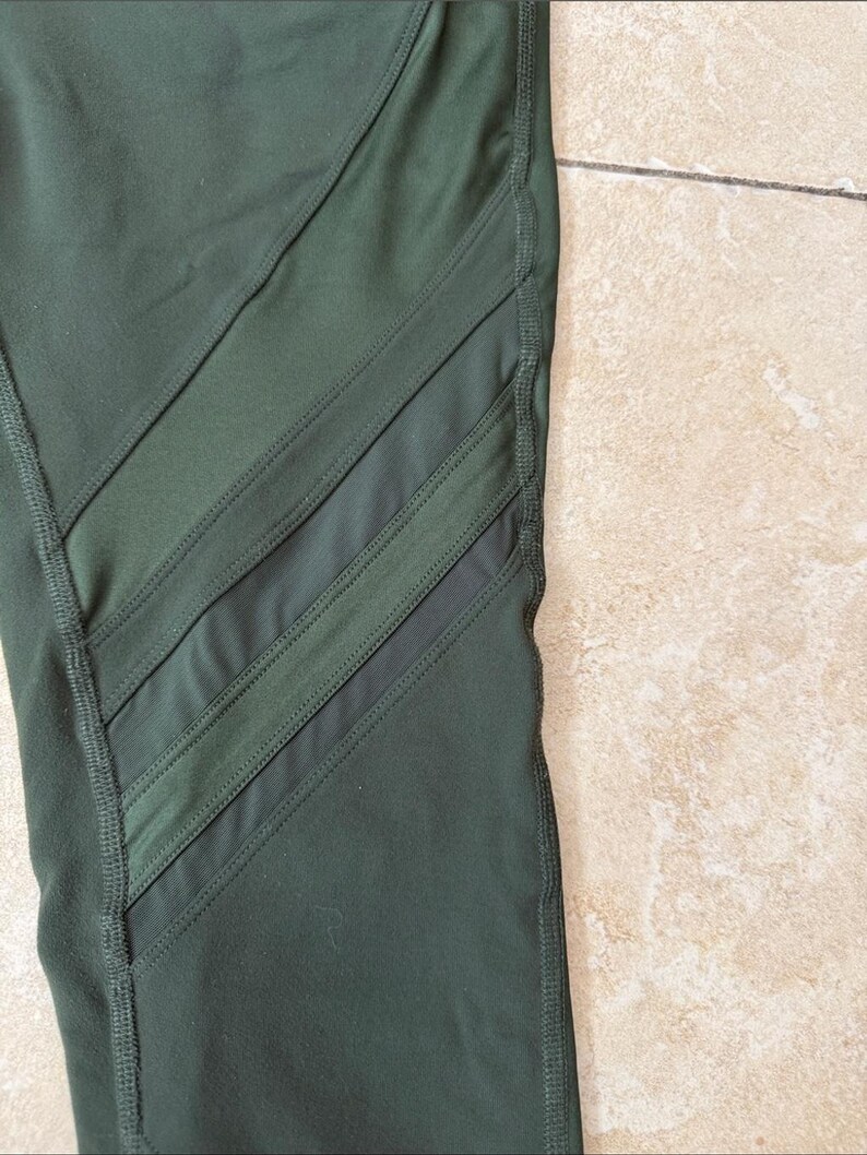 Peut inclure: Legging vert olive fonc&eacute; dot&eacute; de trois panneaux en maille diagonaux sur le c&ocirc;t&eacute;. Le v&ecirc;tement est con&ccedil;u avec des coutures renforc&eacute;es visibles le long des jointures et des bords des inserts en maille transparente, cr&eacute;ant un aspect athl&eacute;tique structur&eacute;.