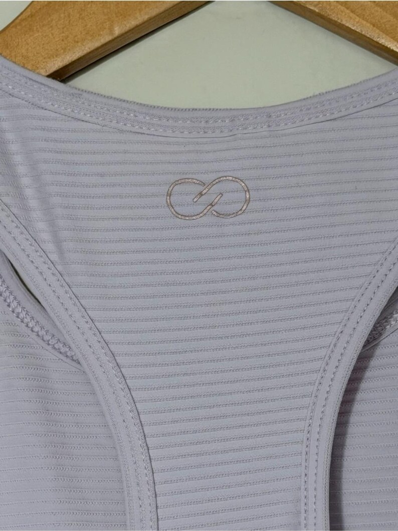 Peut inclure: Un d&eacute;bardeur dos nageur lilas clair pr&eacute;sentant une subtile texture c&ocirc;tel&eacute;e horizontale sur tout le tissu. Au centre de la poitrine se trouve un petit logo brod&eacute; en forme d'infini, r&eacute;alis&eacute; avec un fil lilas clair assorti.