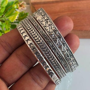 May include: A set of four silver-toned metal bangles featuring distinct embossed patterns. The designs include a row of hearts, a repeating leaf or wheat motif, a geometric linear pattern, and a series of small bird figures in flight.