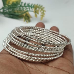 Puede incluir: Un conjunto de múltiples brazaletes de color plateado, cada uno con una textura trenzada similar a una cuerda. Los brazaletes son circulares, metálicos y reflectantes, apilados para formar un grupo coherente de piezas de joyería.