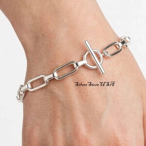 Op de afbeelding: Een zilveren schakelarmband met een knevelsluiting. De armband bestaat uit gepolijste, langwerpige ovale schakels die in een reeks met elkaar verbonden zijn. De tekst Silver Store USA staat in een zwart cursief lettertype bij de sluiting.