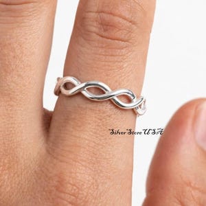 Puede incluir: Un anillo de plata pulida con un diseño trenzado o de infinito continuo que envuelve el dedo. El metal tiene una superficie lisa y reflectante. El texto Silver Store USA es visible en la esquina inferior derecha de la imagen.