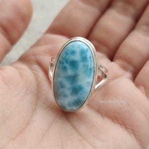 May include: A sterling silver ring featuring a large, oval-shaped, polished blue Larimar gemstone with a distinct white, cloud-like pattern. The stone is set in a smooth metal bezel, and the band consists of a split, polished silver shank.