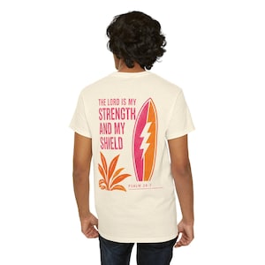 Könnte beinhalten: Ein cremefarbenes Kurzarm-T-Shirt mit einem Grafikdruck auf der Rückseite. Das Design zeigt den Text "THE LORD IS MY STRENGTH AND MY SHIELD" in Pink, ein pink-orangefarbenes Surfbrett mit einem weißen Blitz und darunter "PSALM 28:7".
