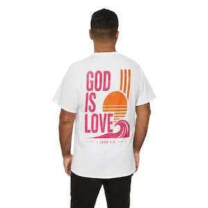Könnte beinhalten: Ein weißes Kurzarm-T-Shirt mit einem Grafikdruck auf der Rückseite, der den Text GOD IS LOVE und 1 JOHN 4:8 zeigt. Das Design enthält eine stilisierte orangefarbene Sonne mit vertikalen Streifen und eine rosa Meereswelle unter dem Schriftzug.