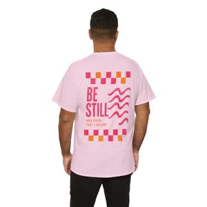 Könnte beinhalten: Ein hellrosa T-Shirt mit kurzen Ärmeln und einem Grafikdruck auf der Rückseite, der von einem rosa-orangen Karomuster umrahmt ist. Der Text BE STILL AND KNOW THAT I AM GOD steht in Rosa und Orange neben drei rosa Wellenlinien.
