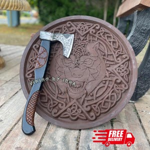 May include: A hand axe with a black handle featuring brown textured grip sections and a metal head engraved with intricate patterns, resting against a circular brown wooden plaque carved with a wolf face and detailed Celtic knotwork designs.