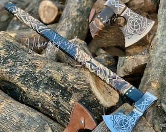 Può includere: Un'ascia a mano con manico in legno intagliato scuro con intricati motivi intrecciati e una testa in acciaio incisa con un design a nodo triquetra. L'ascia include un fodero in pelle marrone e mostra il testo Vikingbyisha sulla lama.