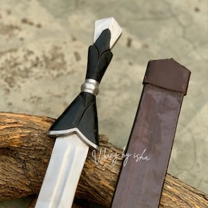 Può includere: Un pugnale con lama in acciaio a doppio taglio, impugnatura nera con accenti color argento e pomolo a forma di punta stilizzata. Il pugnale è accompagnato da un fodero marrone. Il testo "Viking by isha" è visibile sulla lama.