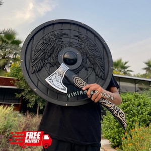 May include: A round black shield featuring a central embossed bear head flanked by two ravens, paired with a matching black axe with intricate silver-toned engravings on the blade and handle, marked with the text VIKING BY ISHA.